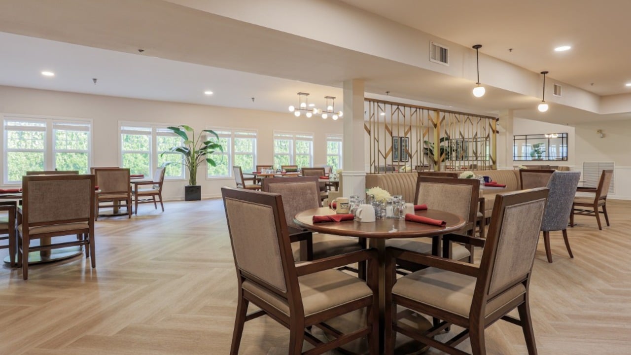A spacious, well-lit dining area with round tables set for meals, cushioned chairs, large windows, wood flooring, potted plants, and modern light fixtures creating a welcoming atmosphere.