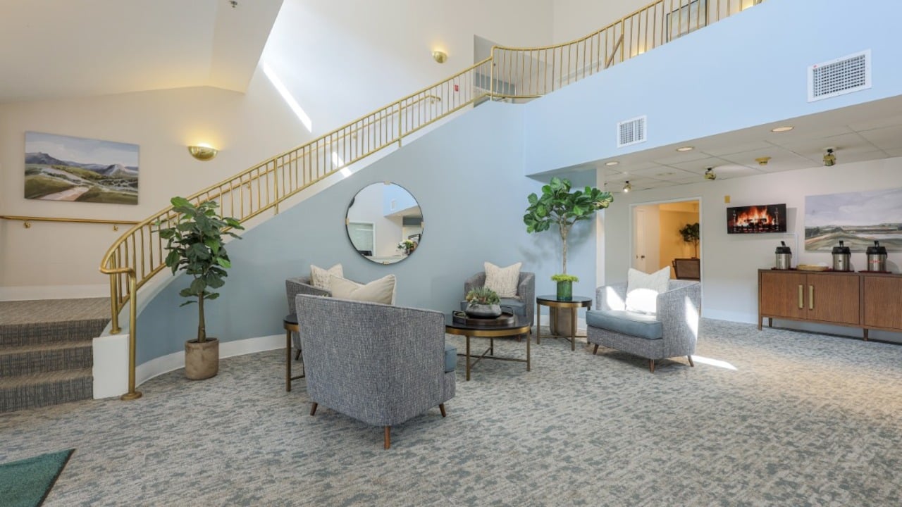 Modern lobby with blue walls, a round mirror, two gray chairs, a loveseat, plants, and a gold staircase. Wall art and a cabinet with decor complete the bright, welcoming space.