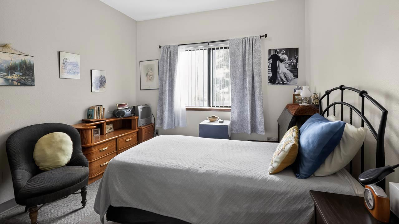 A cozy bedroom with a neatly made bed, a cushioned chair, wooden dresser, books, framed art, and a window with sheer curtains letting in daylight. Black-and-white photos hang on the wall.