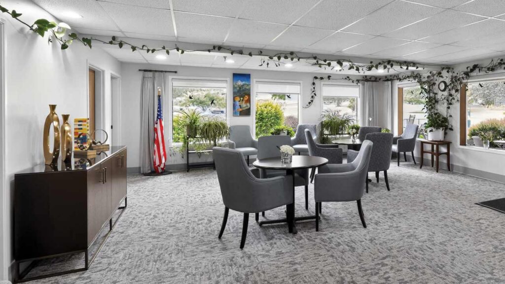 A bright, modern lounge with gray armchairs around tables, large windows, plants, an American flag, and decorative items on a sideboard.