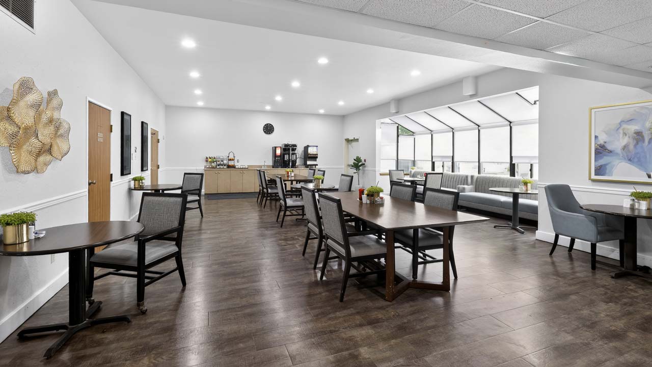 Bright, modern dining area with dark wood floors, tables, chairs, a coffee station, wall art, and large windows letting in natural light. Small plants decorate the tables.
