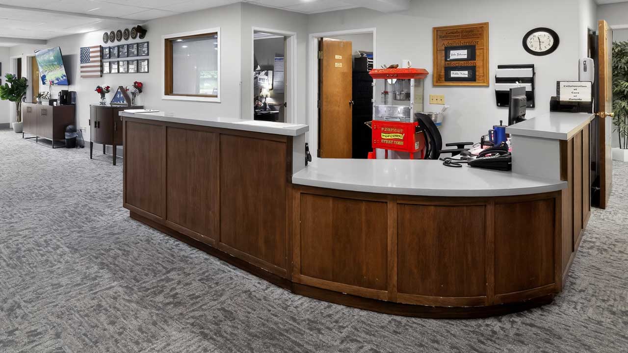 Reception area with curved wooden desk, office equipment, red popcorn machine, wall clock, and framed items on light-colored walls; gray carpeted floor and adjacent hallway with cabinets and decor.