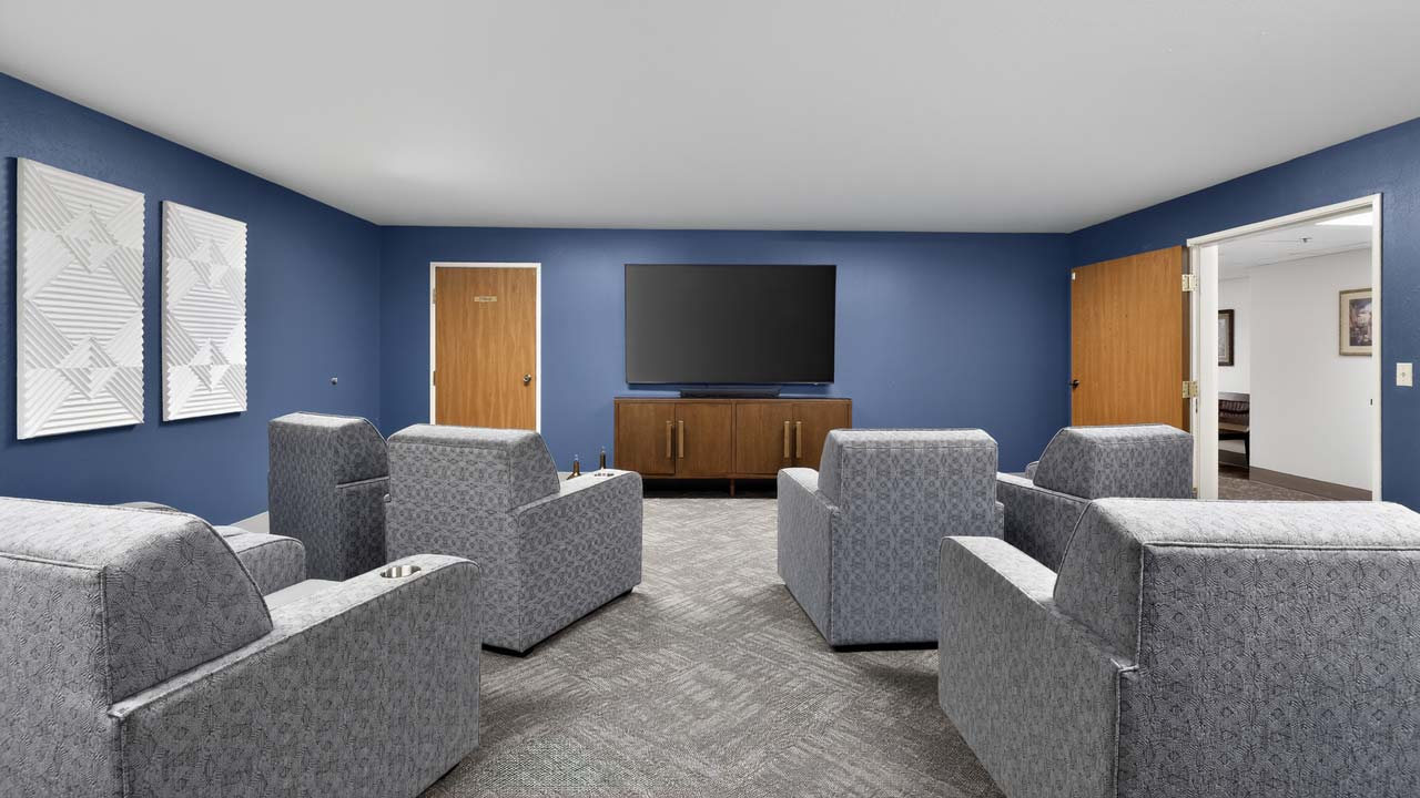 A modern lounge with four gray armchairs facing a large flat-screen TV on a wooden console, blue accent walls, and abstract art. Two closed doors and one open door are visible.