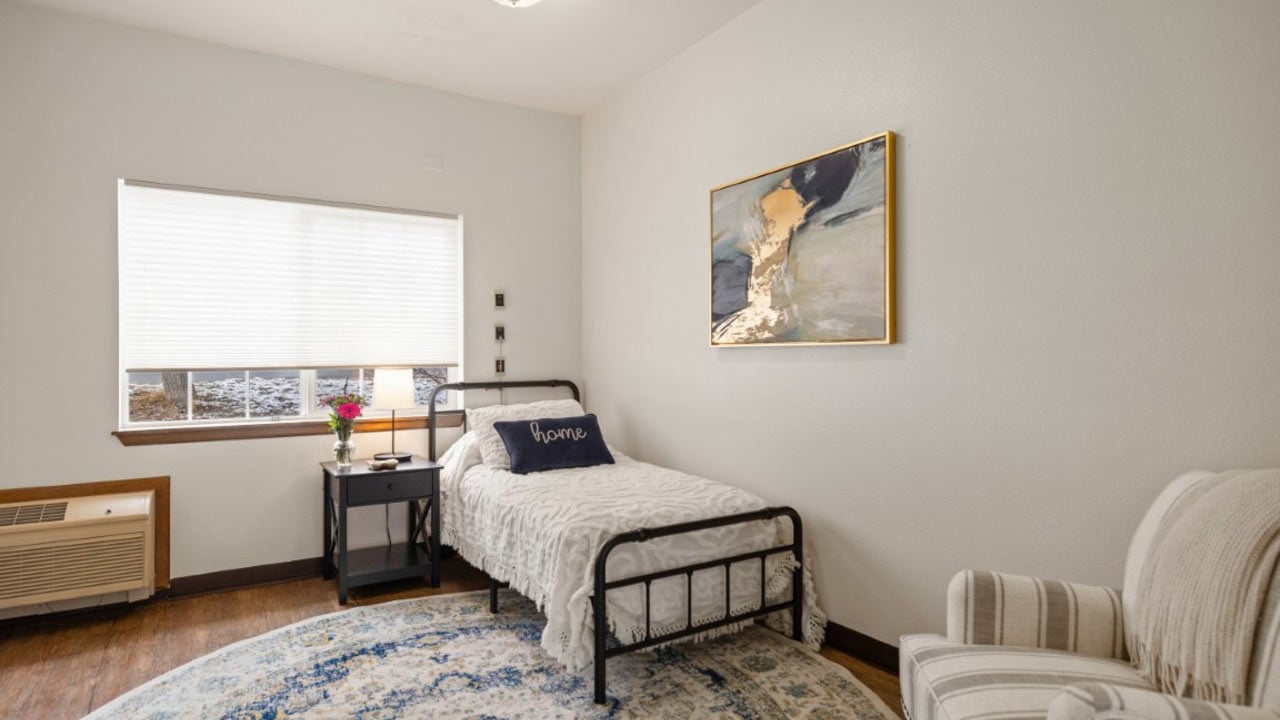 A cozy bedroom with a single bed, “Home” pillow, nightstand with a lamp and flowers, armchair, abstract wall art, window with blinds, and an air conditioning unit.