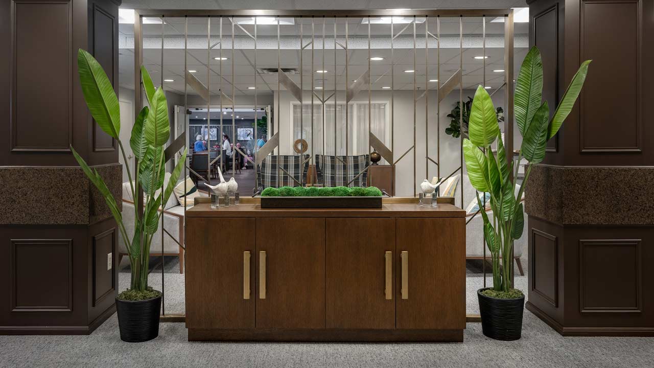 Modern lobby with a wooden cabinet, two tall potted plants, geometric mirrored wall, and decorative items. People are visible in the background through an open area.