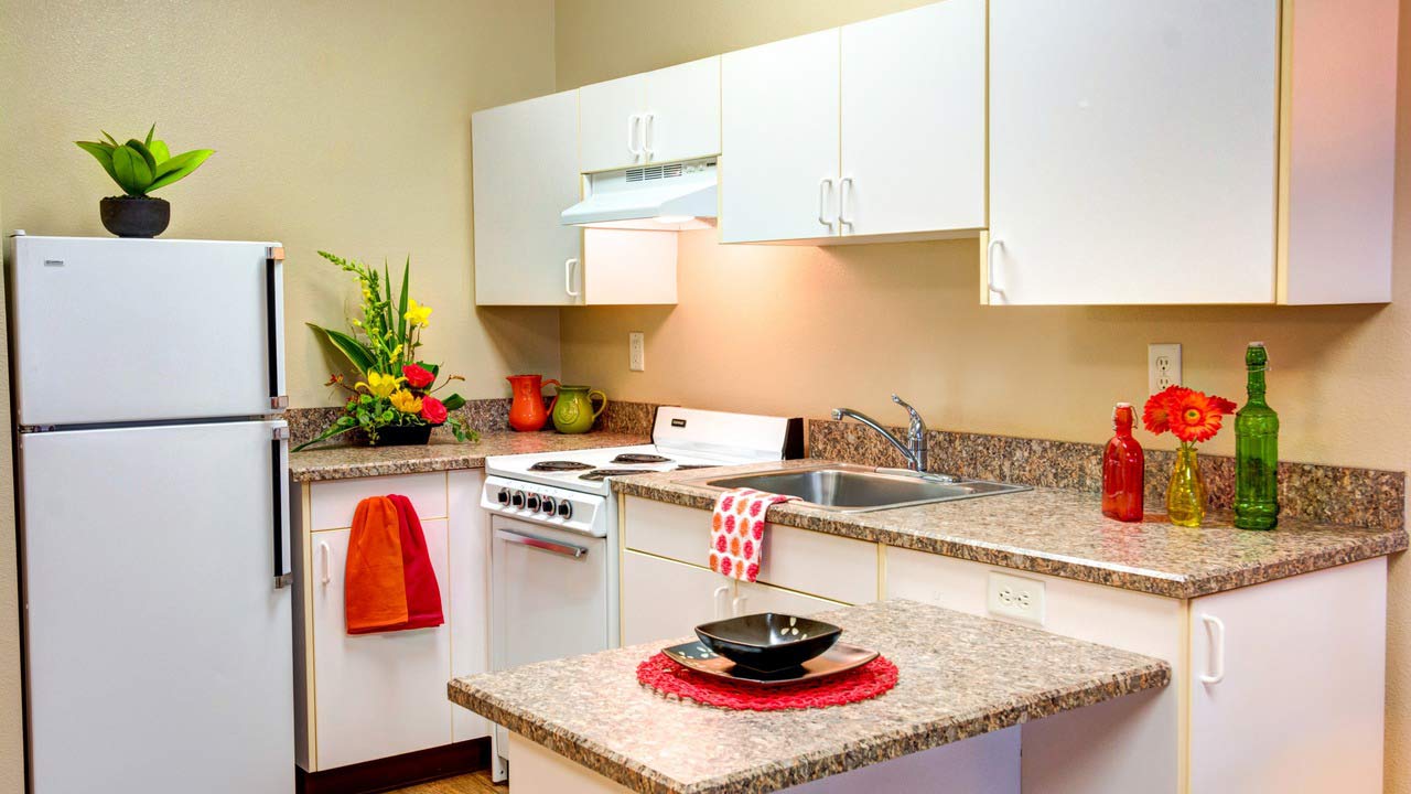 A small kitchen with white cabinets, a white fridge and stove, granite countertops, colorful flowers, red and green decor, and a sink. A black bowl and plate sit on a red placemat on the counter.