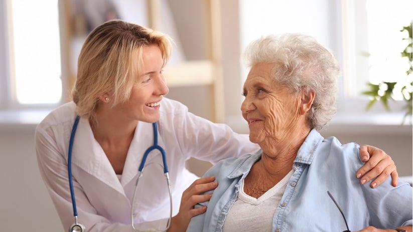A healthcare provider with a stethoscope smiles and talks to an elderly woman, gently touching her shoulder, in a bright, comfortable room, demonstrating compassionate health management in senior living.