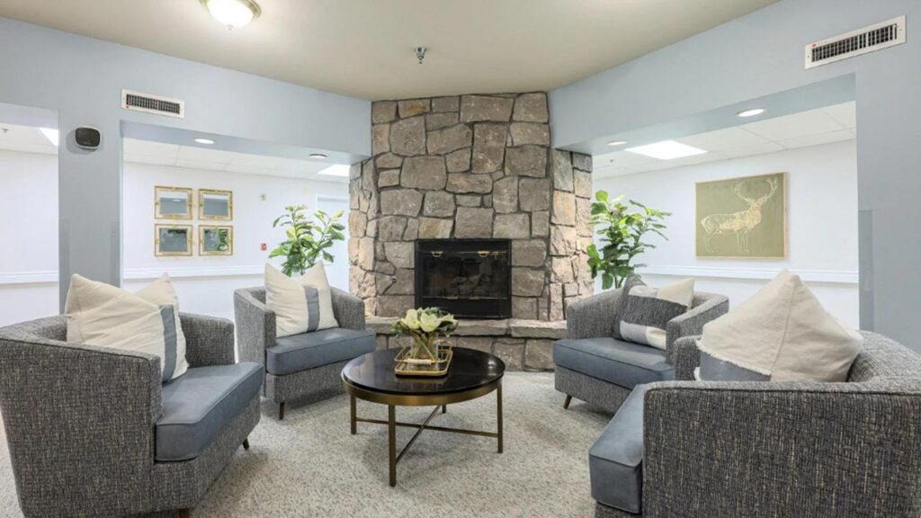 Four gray armchairs with white pillows are arranged around a round coffee table in front of a stone fireplace, creating an inviting assisted living space with green plants and framed artwork brightening the room.
