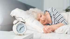 A white analog alarm clock sits on a bedside table in front of an older person sleeping, illustrating the importance of sleep hygiene for seniors.
